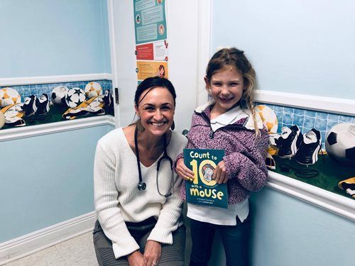 Woman and young girl holding a colorful “Count to Mouse” book in a bright room with penguin wall border