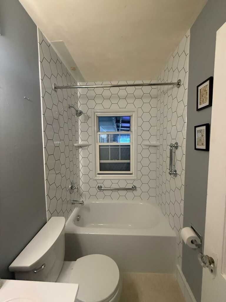 Small bathroom with white hexagon tile shower, toilet, and bath, next to a gray wall.