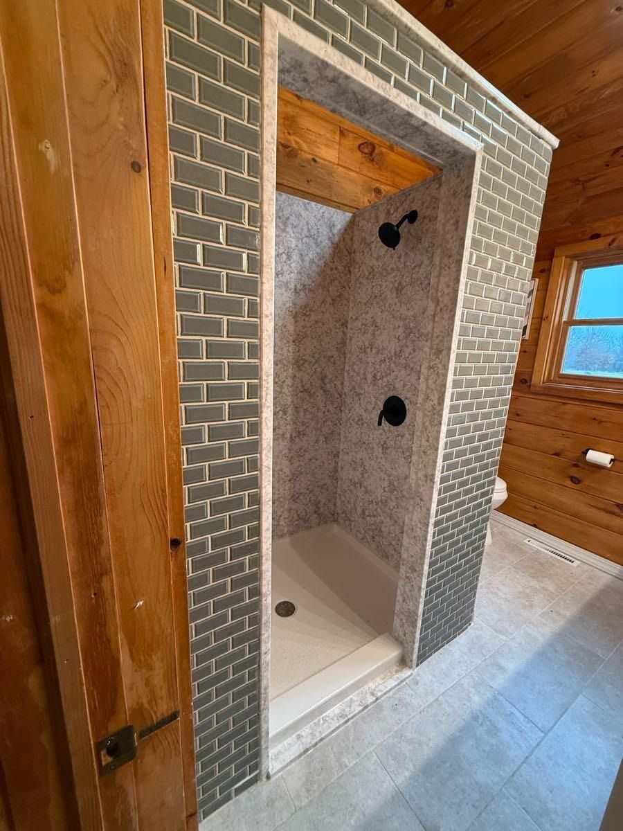 Shower stall with gray brick-like tile surround and white floor, built in a wood-paneled bathroom.