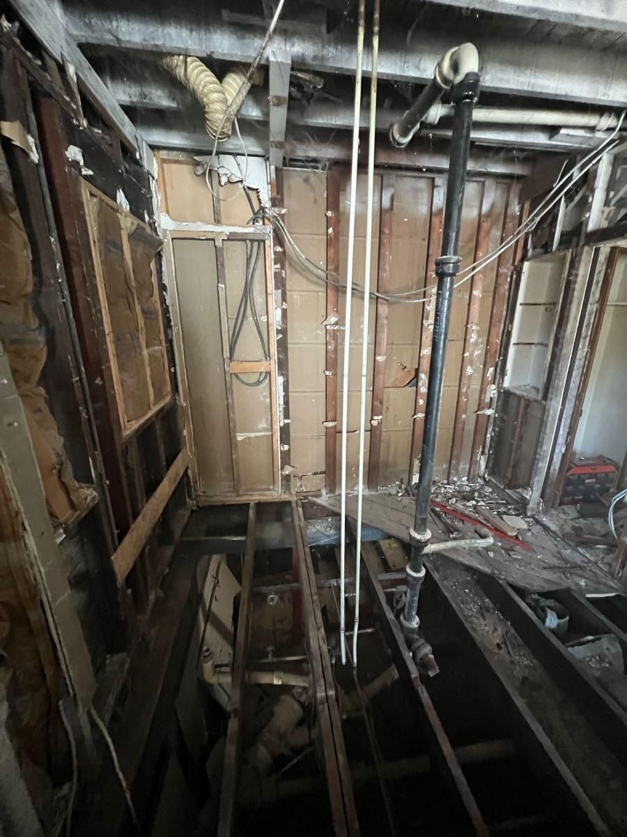 Interior demolition. Exposed beams, pipes, and wood framing. Debris on floor.