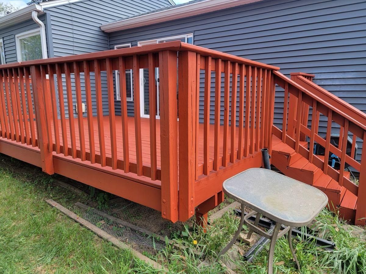 Red painted wooden deck and steps attached to a gray house, with a small table on the grass.