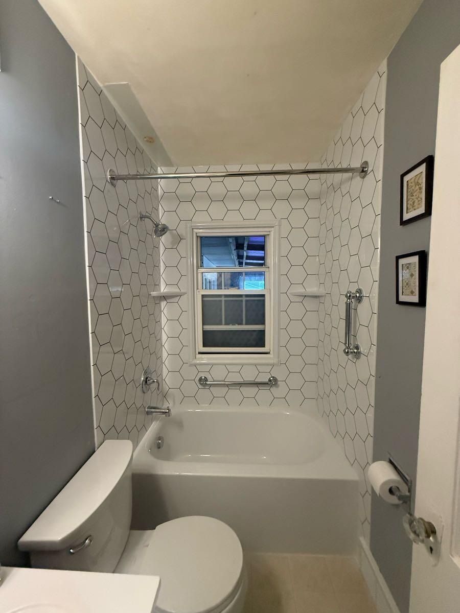 Small bathroom with hexagonal tiled shower, bathtub, toilet, and window.