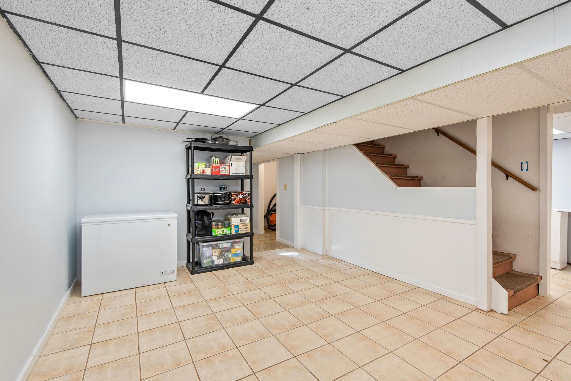Basement storage area with tile floor, freezer, shelving unit, and stairs leading up.