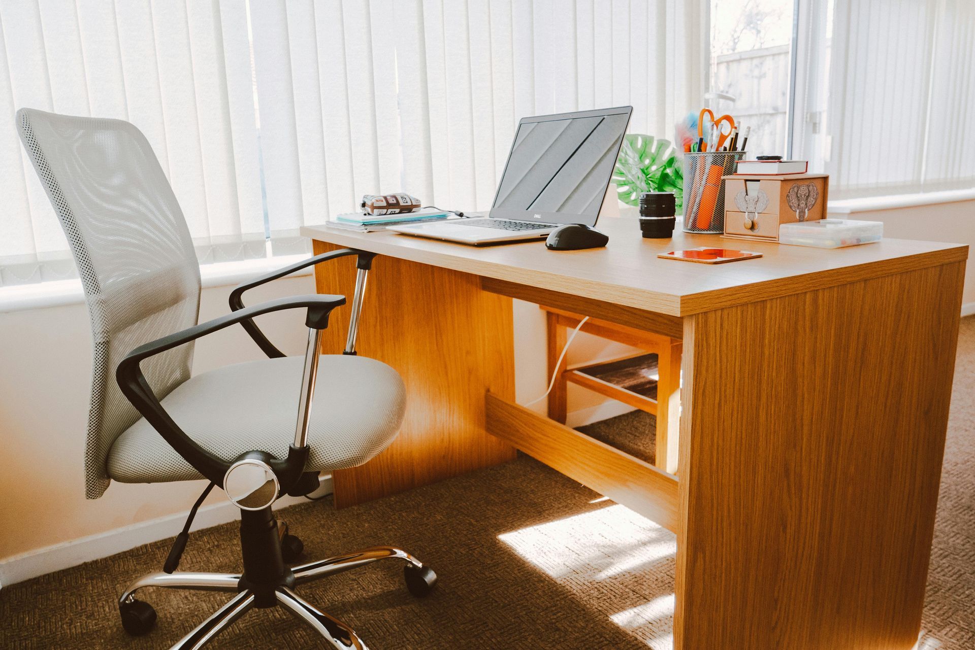 Office desk with laptop, chair, and accessories. Sunlight streams through window blinds.