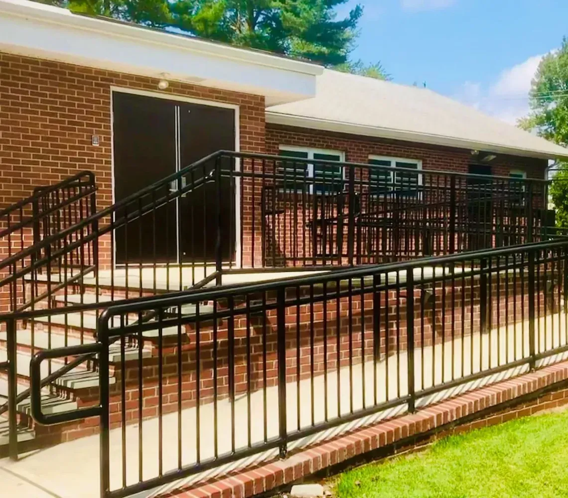 Brick building entrance with black metal ramps and railings, leading to a patio and lawn.