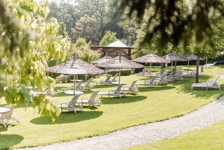 Gartenstühle unter Strohschirmen auf einer Rasenfläche mit einem Weg, Bäumen und einem hölzernen Pavillon im Hintergrund.