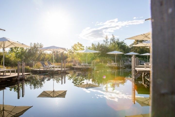 Sonnenbeschienener Pool mit Sonnenschirmen, in denen sich der Himmel spiegelt. Holzstege mit Liegestühlen, umgeben von Grün.