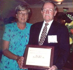Two people in formal attire hold a framed award certificate at a banquet.