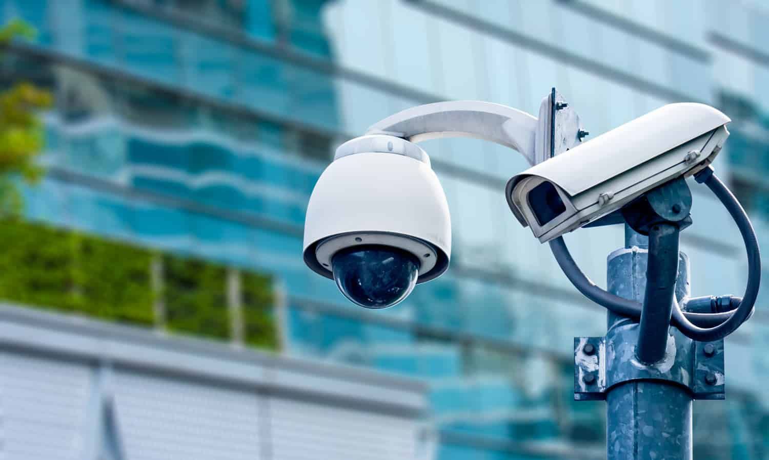 Two white security cameras mounted on a pole outside a glass office building