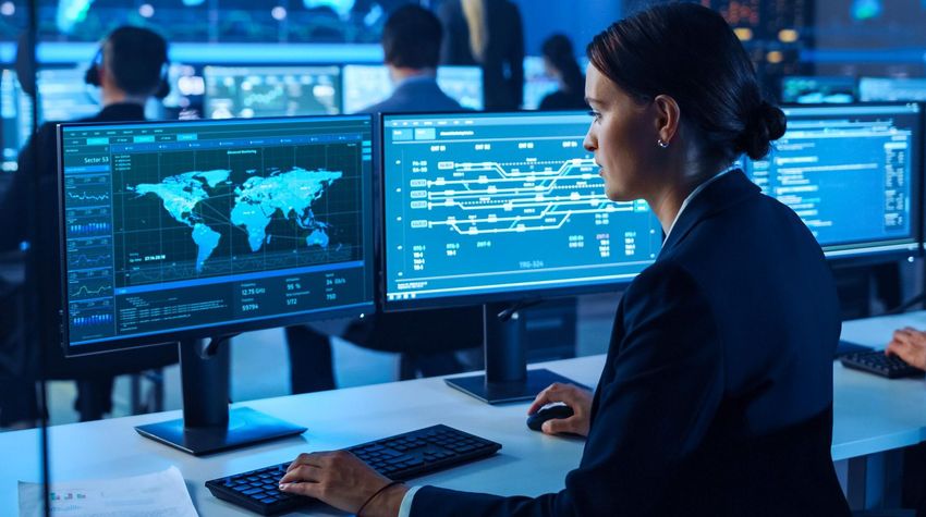 Analyst at a cybersecurity control room with multiple monitors showing world maps and network data charts