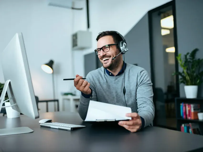 Office worker wearing a headset smiles at a desk, holding papers and a phone beside a computer.