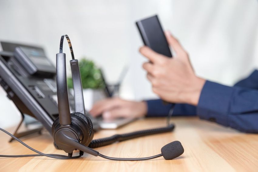 Desk phone headset with microphone beside a person using a smartphone and laptop at an office desk