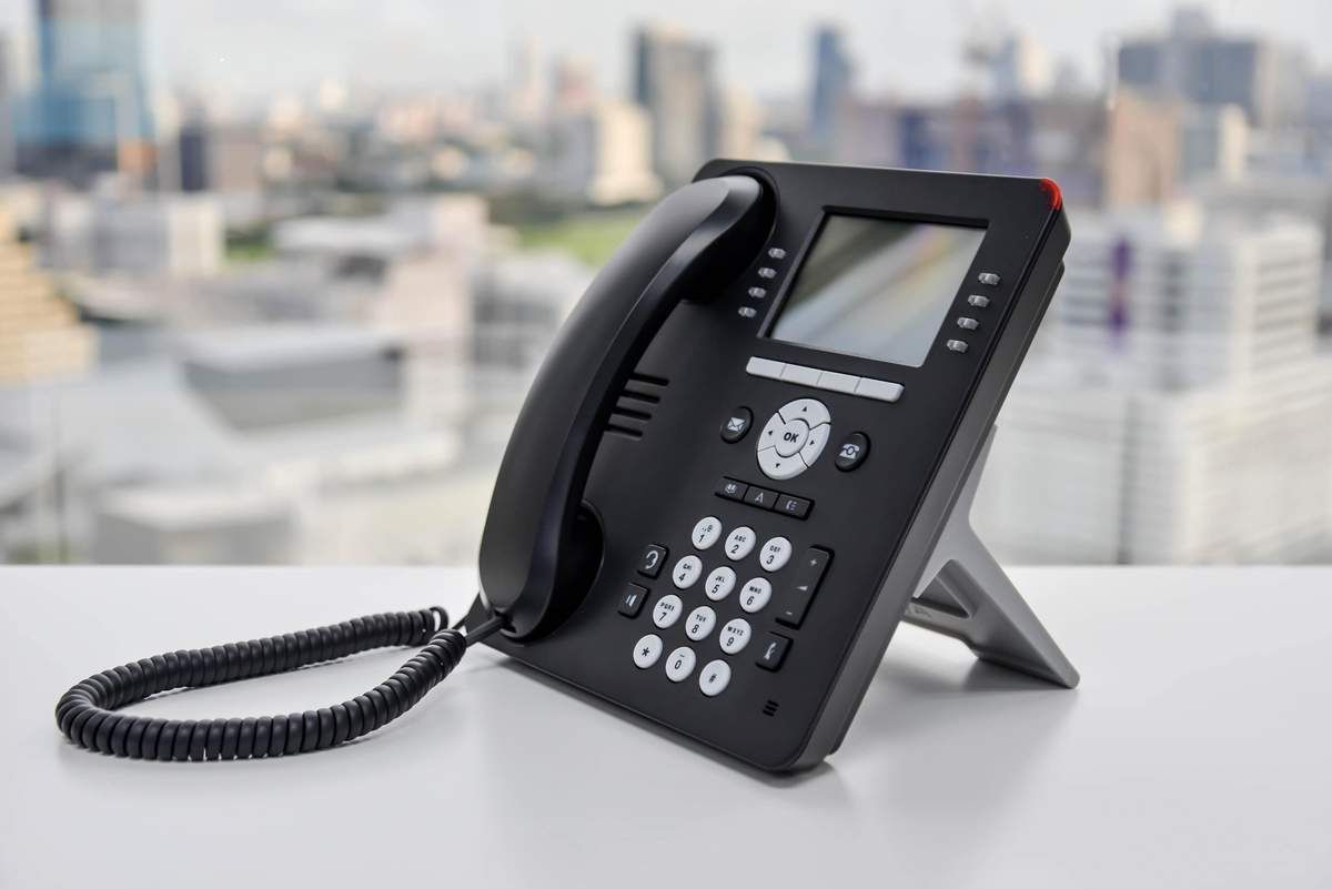 Black office desk phone with handset on a stand, set on a white table with a blurred city skyline behind it