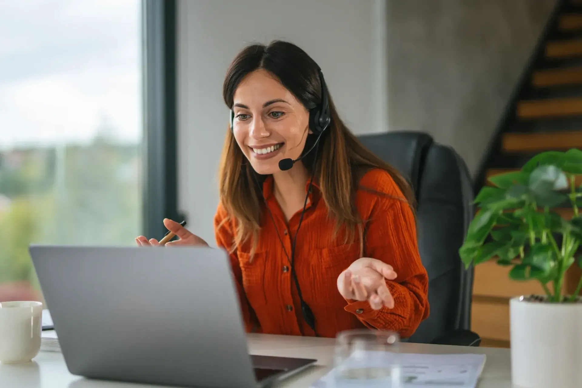 Woman in orange shirt smiling during a video call at a laptop in a bright office