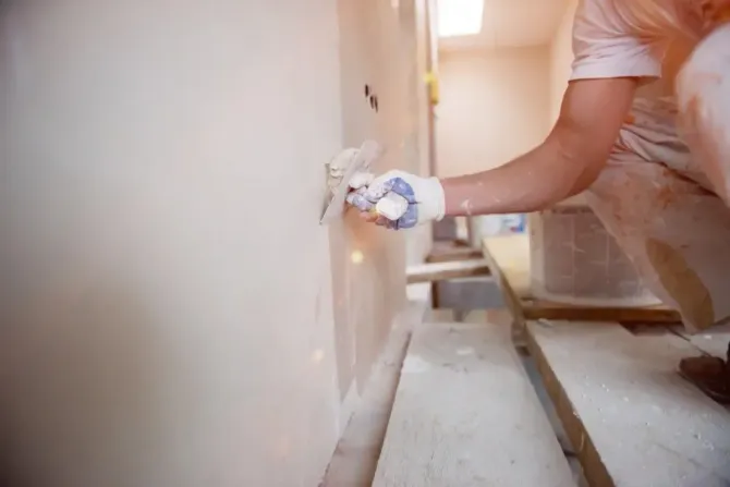 A worker wearing white gloves uses a metal trowel to smooth joint compound over a plaster wall in an indoor space.