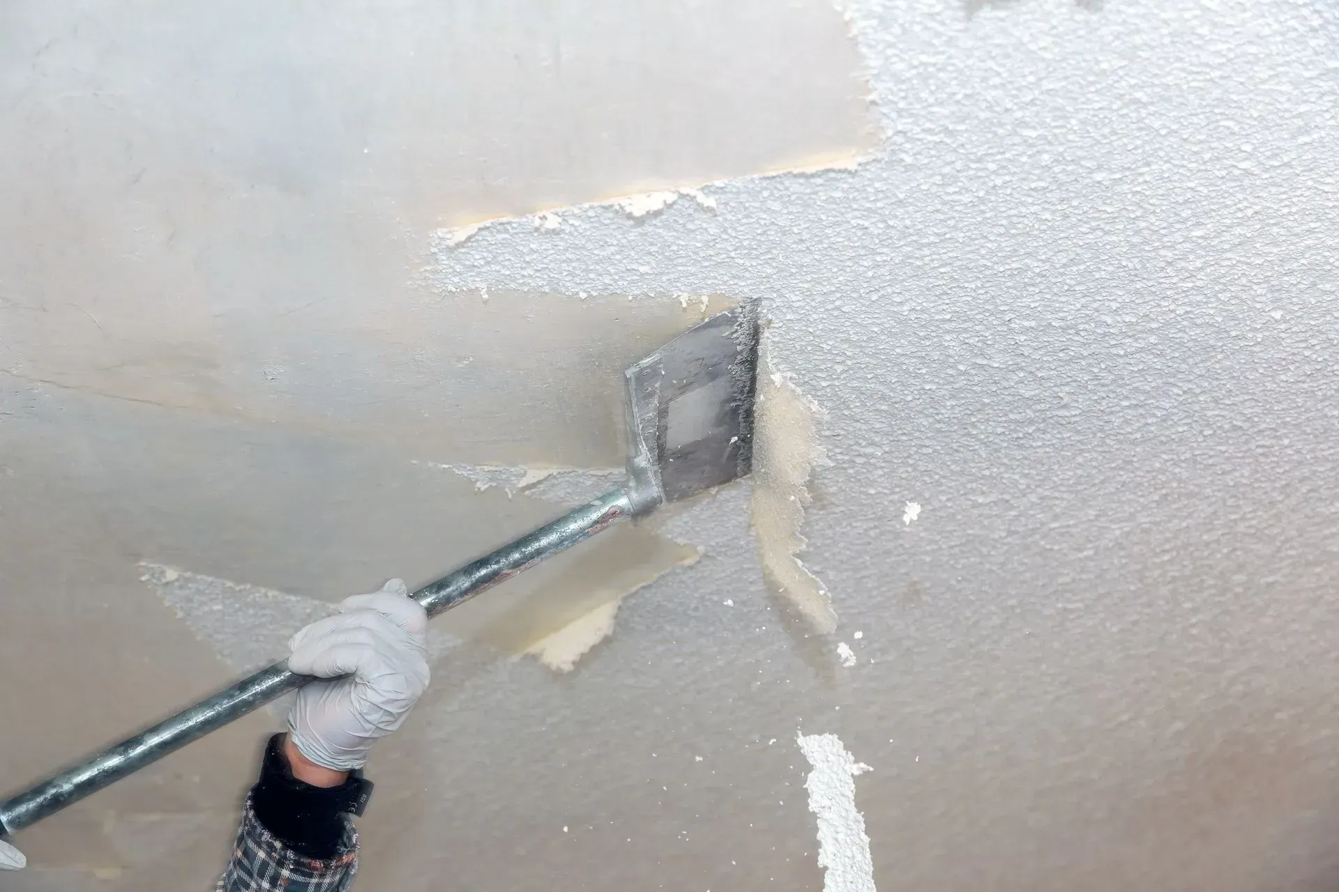 A person wearing a glove scrapes popcorn texture off a ceiling with a long-handled tool.