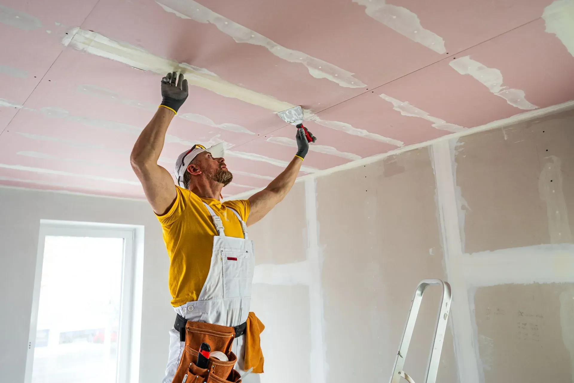 A person wearing a yellow shirt and white overalls applies plaster to a pink drywall ceiling in a room under construction.