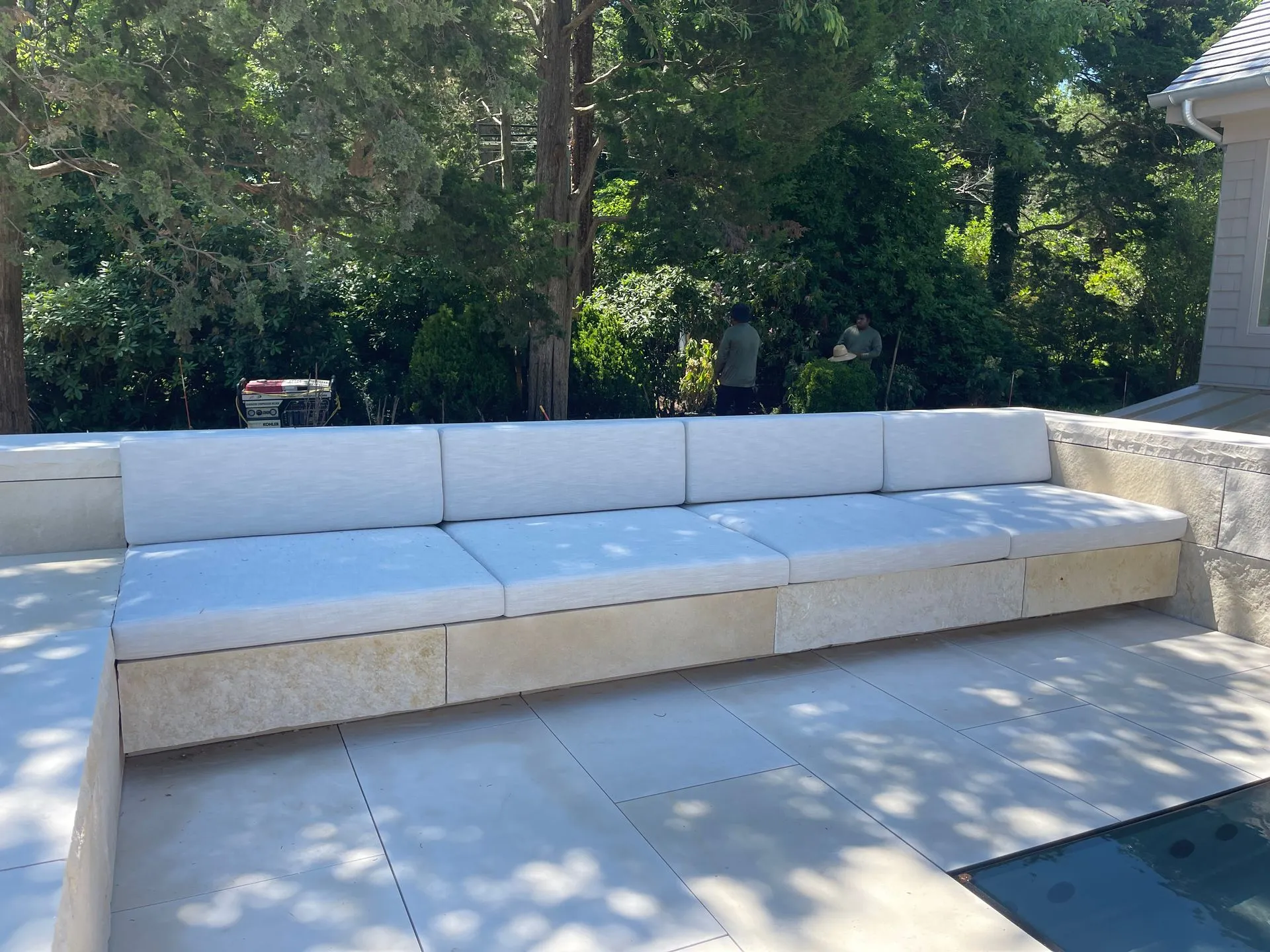 White outdoor sectional sofa with beige cushions on a patio beside a pool, backed by trees and a house.