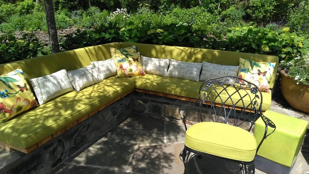Yellow L-shaped outdoor sofa with patterned pillows on a sunlit patio amid greenery