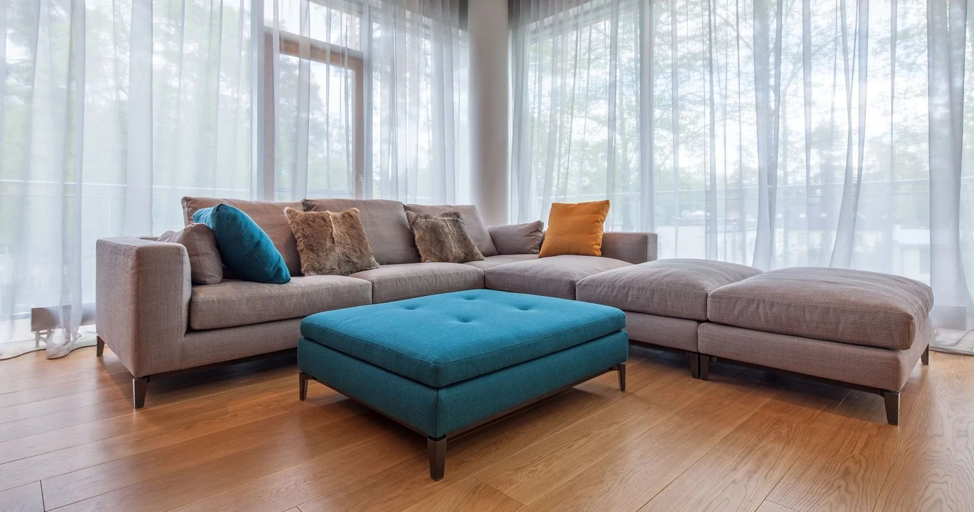 Modern living room with a gray sectional sofa and teal ottoman near bright windows