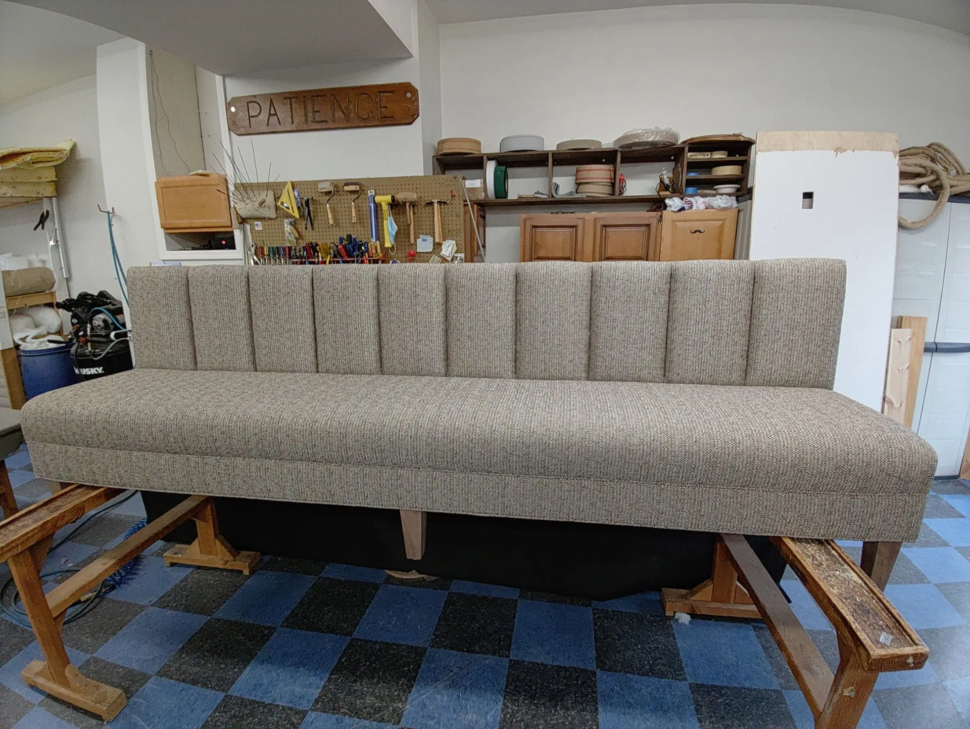 Beige upholstered bench on a workbench in a cluttered workshop with shelves and tools behind it