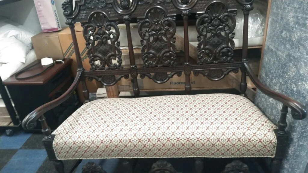 Ornate dark wooden bench with patterned cushion in a room, beside a small side table.