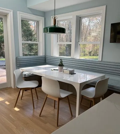 Bright dining nook with white table, four chairs, striped built-in bench, and large corner windows.