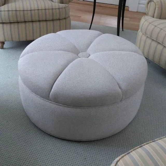 Round light gray tufted ottoman on a carpeted living room floor, with striped chairs nearby