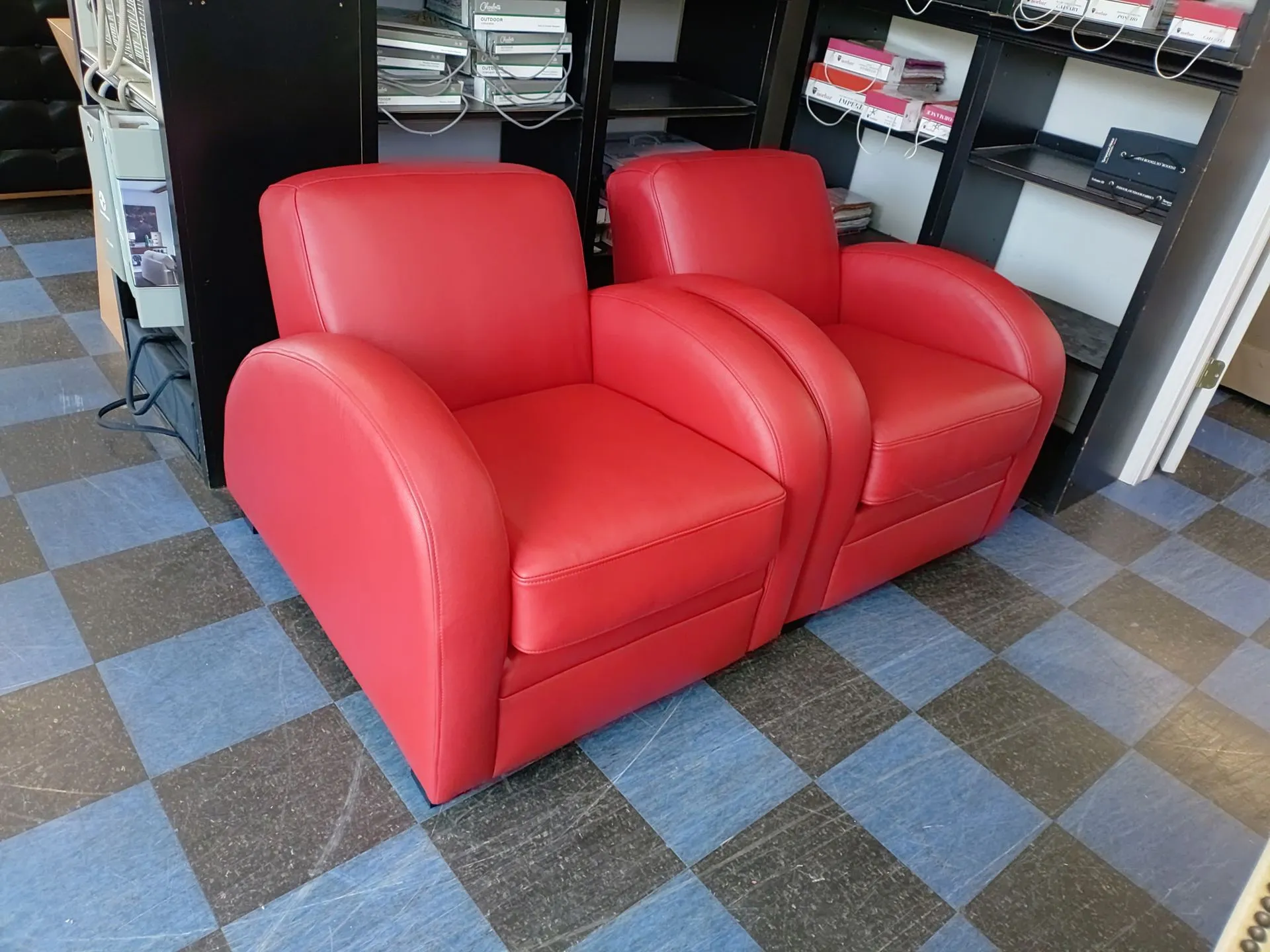 Two red armchairs side by side on a checkered floor in a shop or storage room.