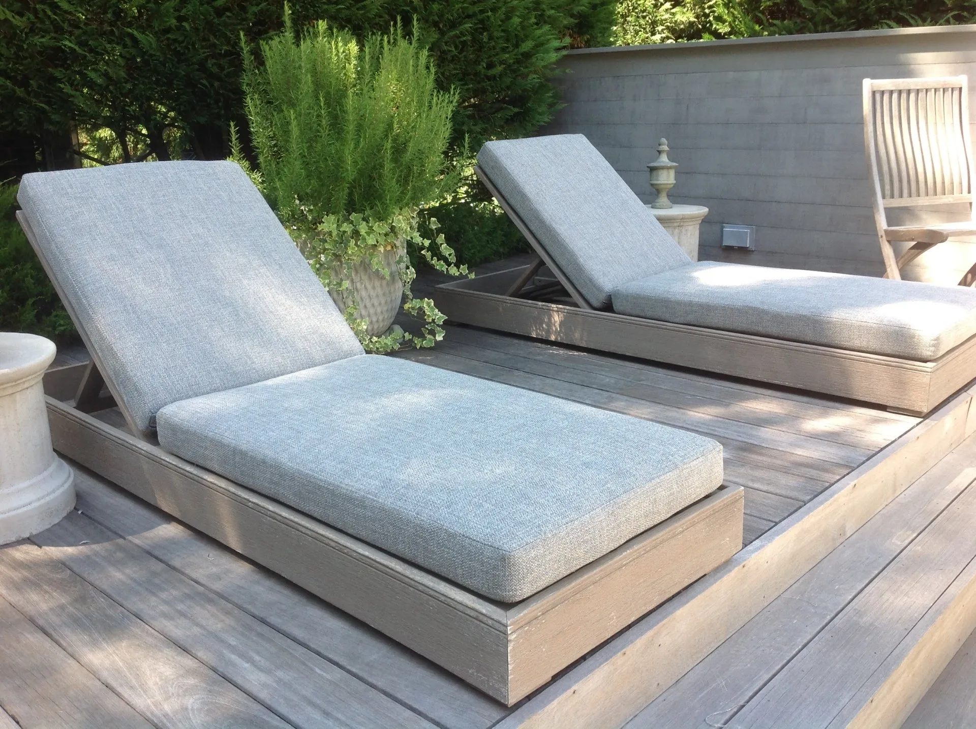 Two light gray outdoor chaise lounges on a wooden deck beside green plants and a gray wall.