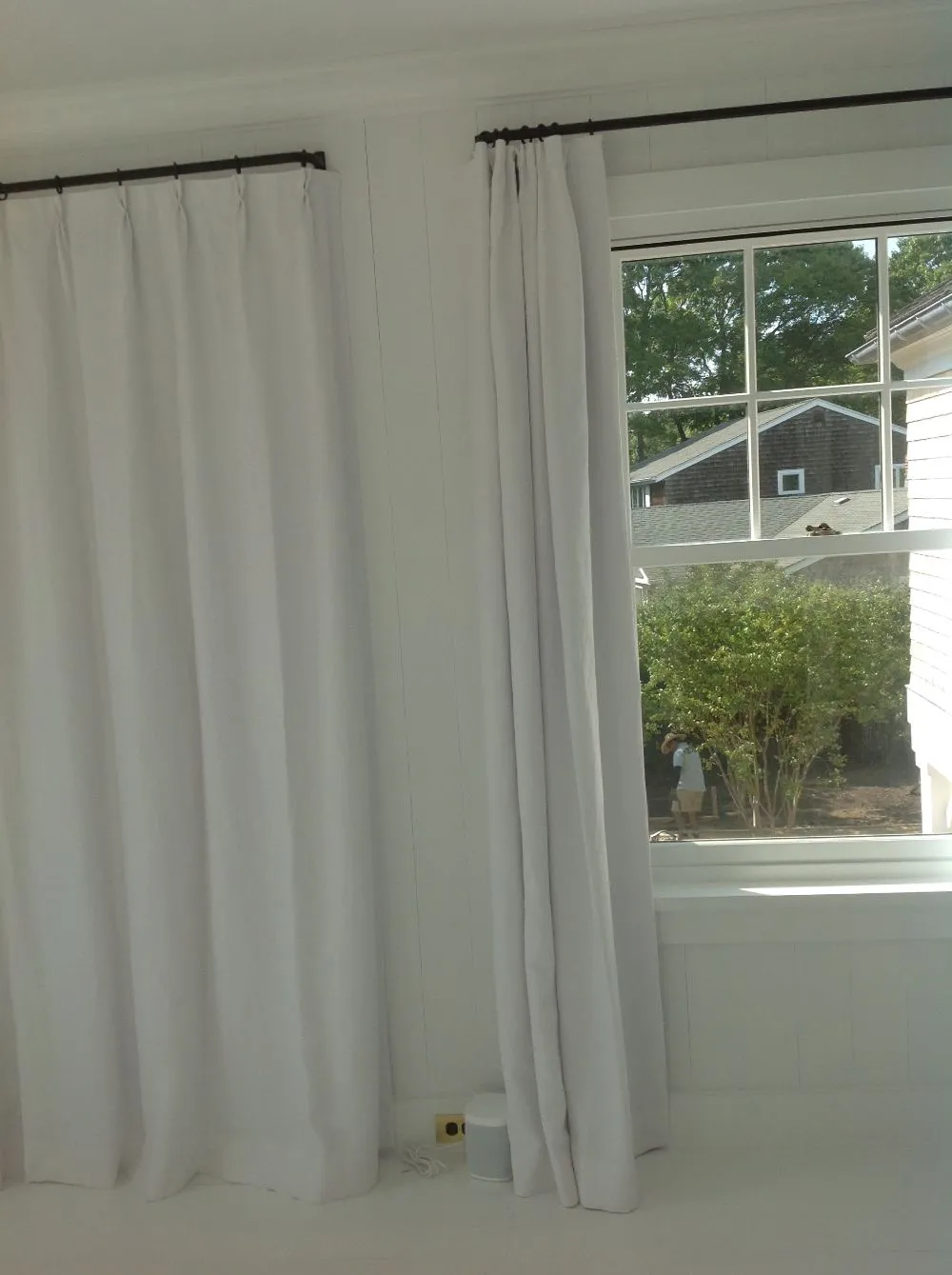 White curtains beside a bright window overlooking green bushes and a neighboring house.