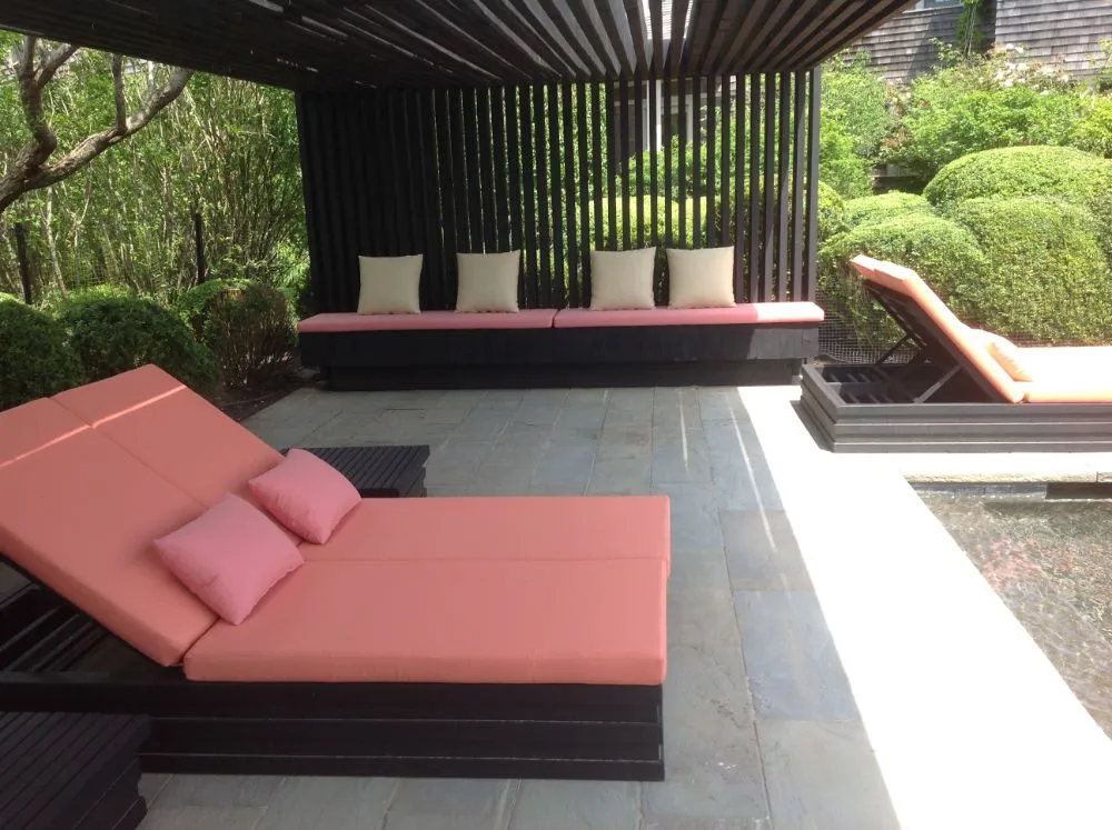 Outdoor lounge with pink cushioned chaise and bench seating under a shaded pergola beside a pool.