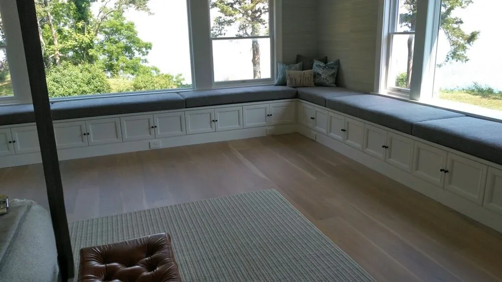 Bright window seat with built-in cabinets, cushions, and a light rug in a sunlit room.