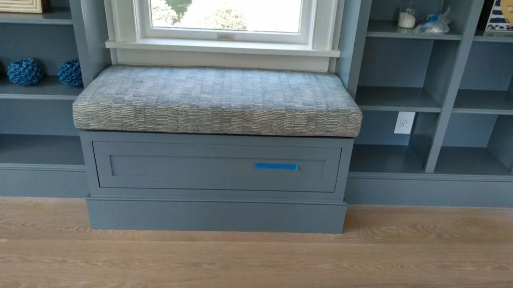 Blue bench with cushion and storage drawers under a window, flanked by built-in shelves.