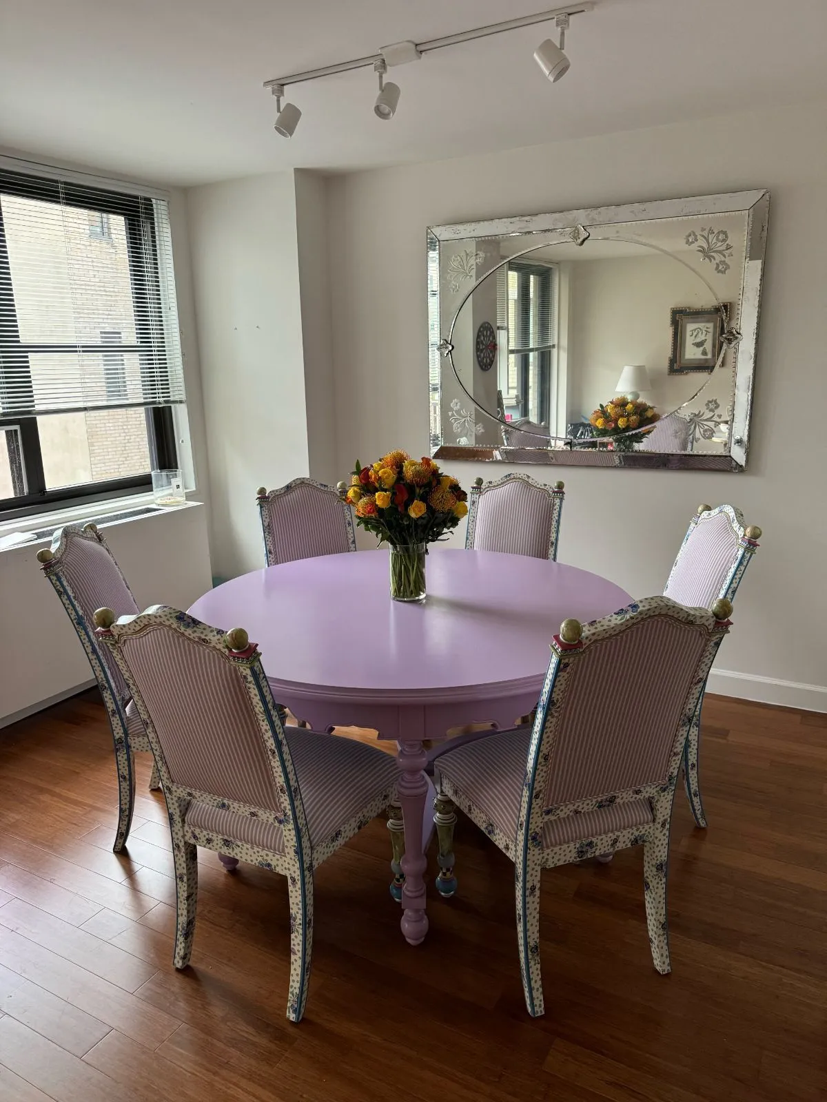 Lavender round dining table with ornate chairs and flowers in a bright room with a mirror and window.
