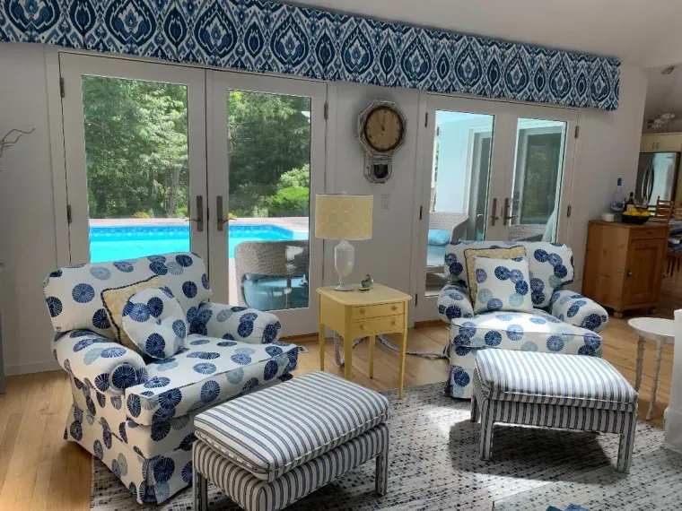 Cozy living room with blue-and-white chairs, ottomans, and a view of a swimming pool through large windows.