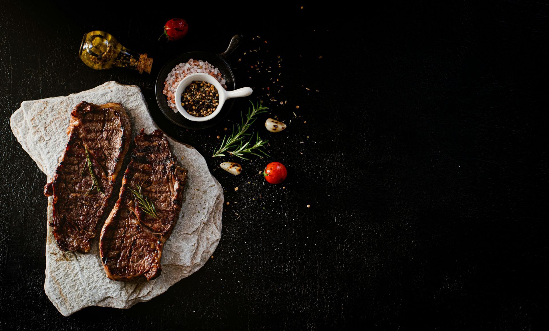 Two cooked steaks on a stone slab, alongside salt, pepper, herbs, tomatoes, and olive oil on a black surface.