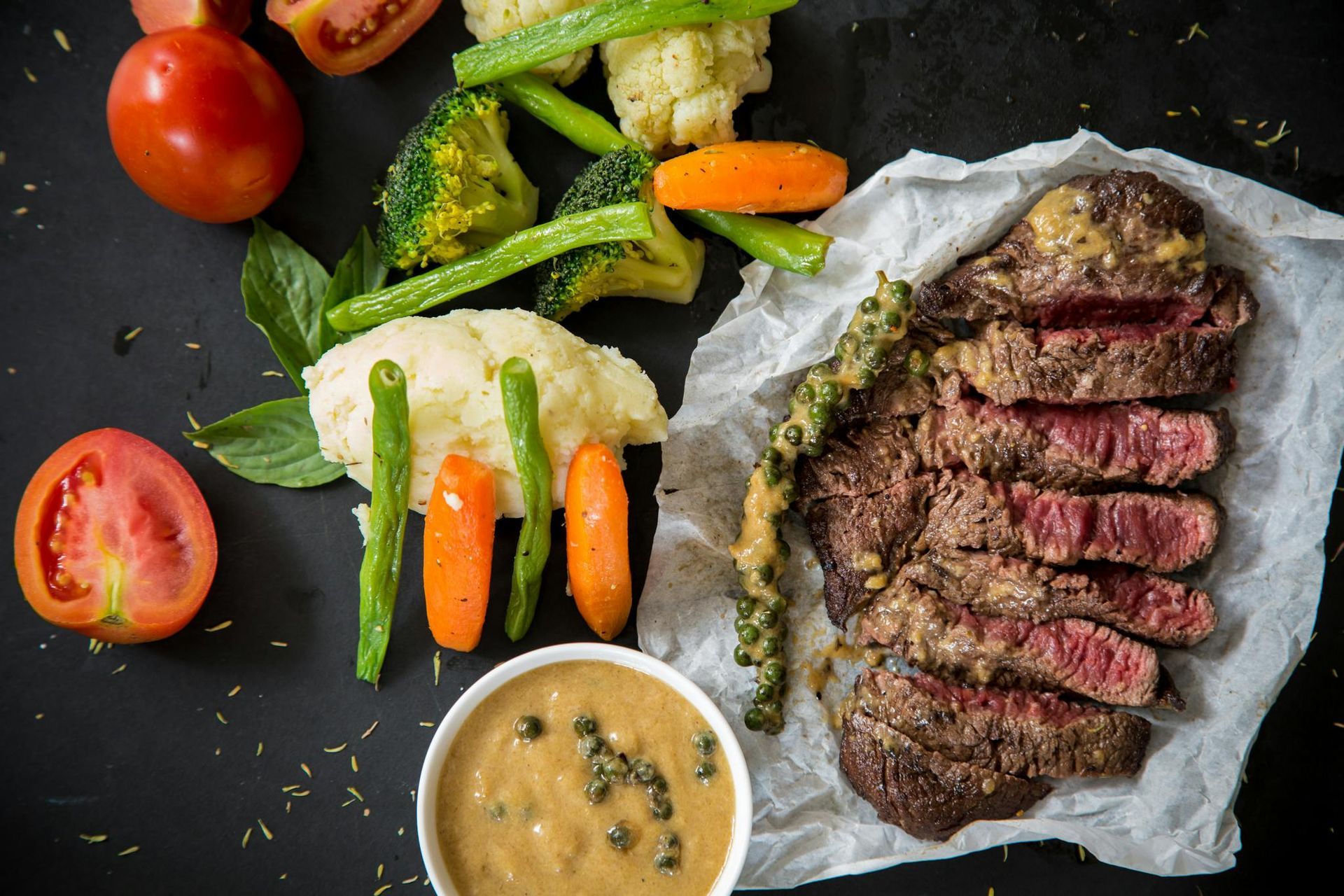 Sliced steak with peppercorn sauce, assorted vegetables, and dipping sauce on a dark surface.
