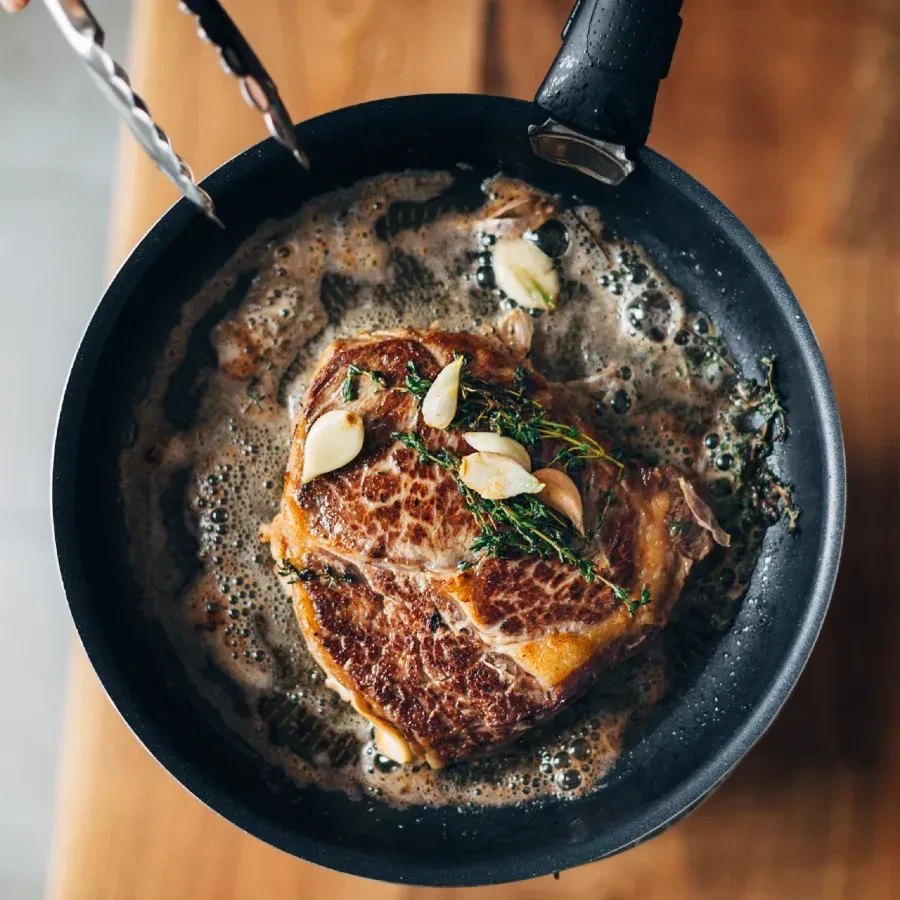 Steak searing in pan with melted butter, garlic, and thyme; tongs reaching in.