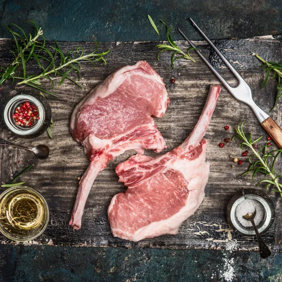 Two raw pork chops with bone-in, on a wooden board with spices, oil, herbs, and a carving fork.
