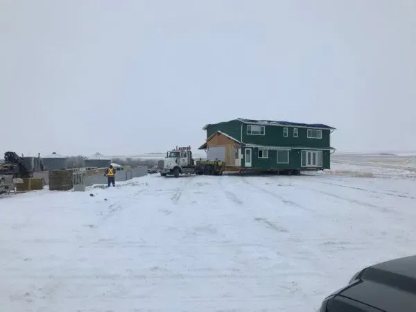 Two-story green house being moved by a truck on a snowy, open field. A person in orange vest stands near.