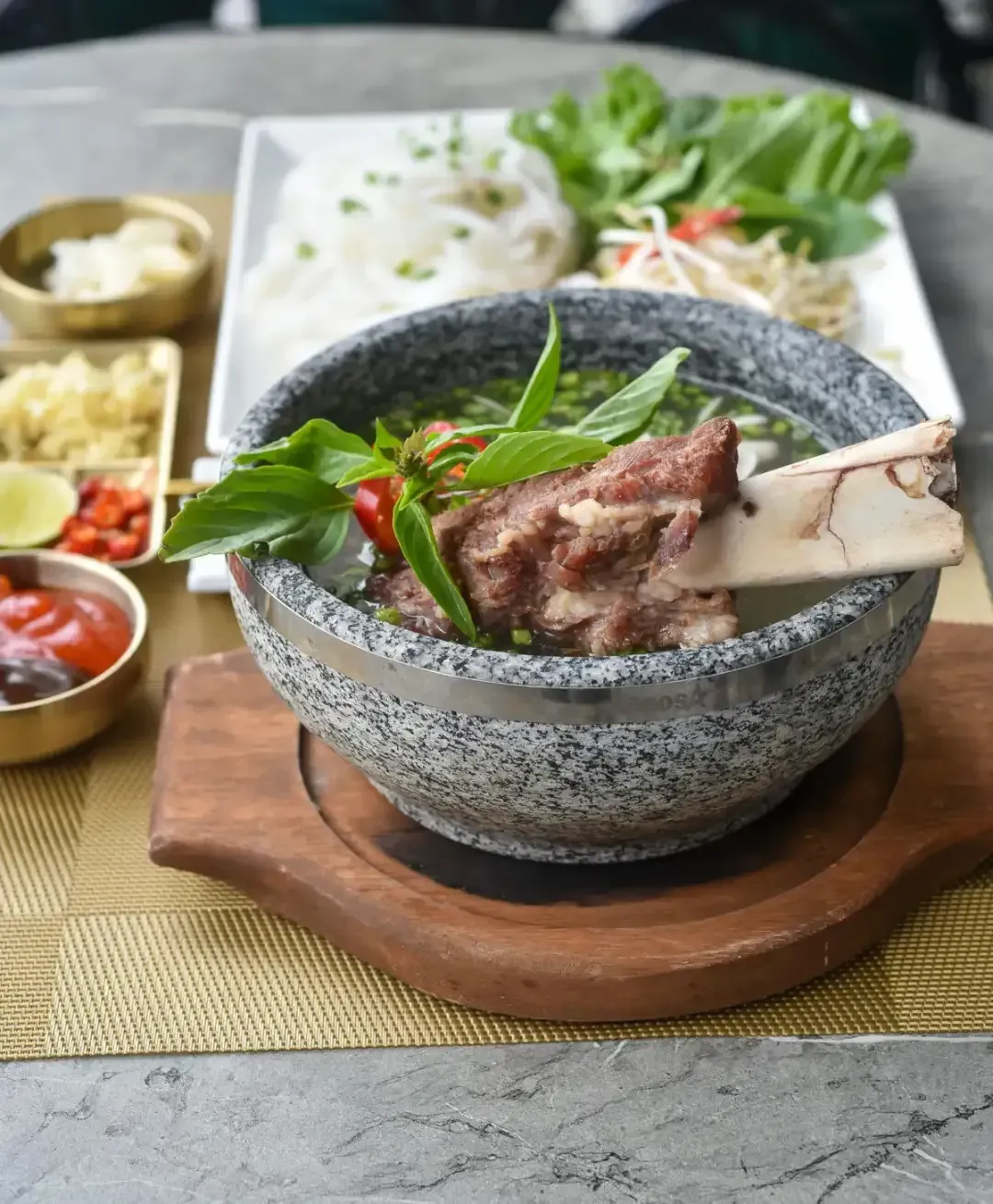 Pho with bone-in short rib in a speckled gray bowl. Side dishes include noodles, herbs, and sauces.