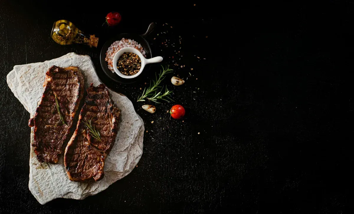 Two cooked steaks on a stone slab, alongside salt, pepper, herbs, tomatoes, and olive oil on a black surface.
