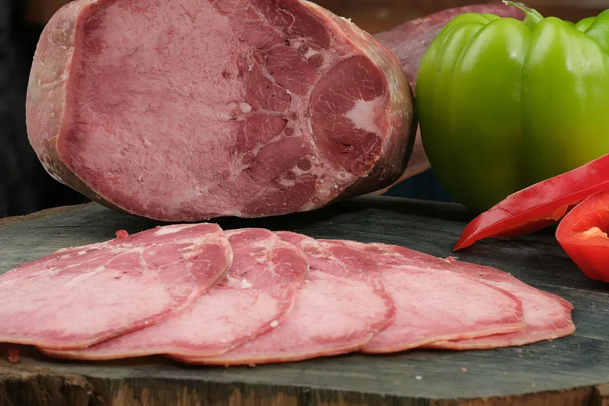 Sliced ham with a whole ham, green pepper, and red pepper on a wooden surface.