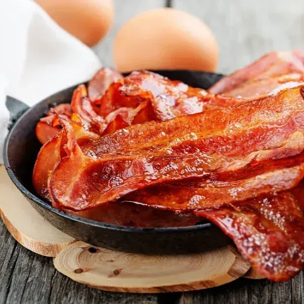 Crispy bacon in a black skillet, two eggs in the background, on a wooden surface.