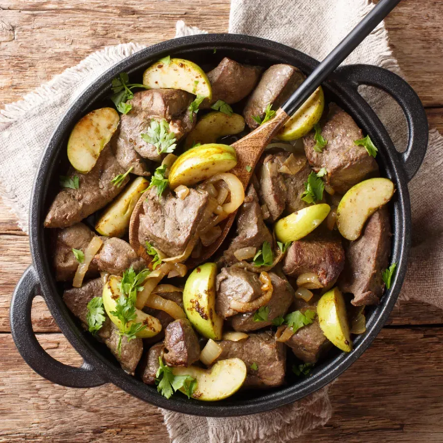 Stewed liver with apples and onions in a black pan with a wooden spoon, on a rustic wooden surface.