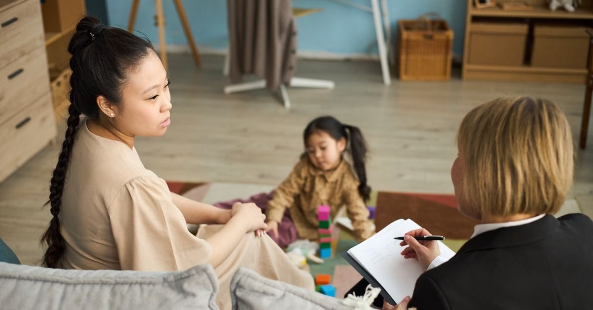 A therapist takes notes while a mother watches her daughter stack colorful blocks during a home session.