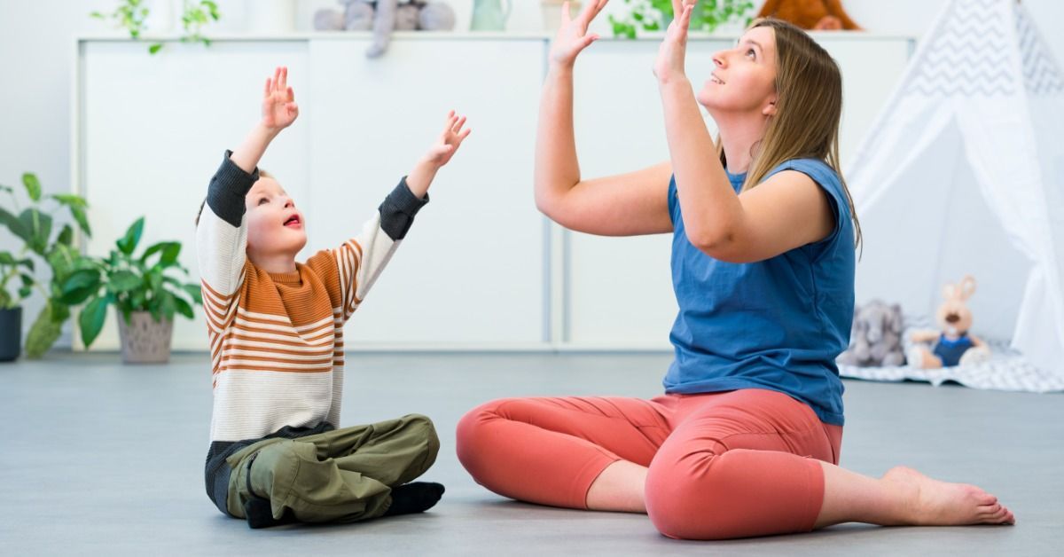 A therapist raises her hands as a young boy copies the movement while they sit together in a bright playroom.