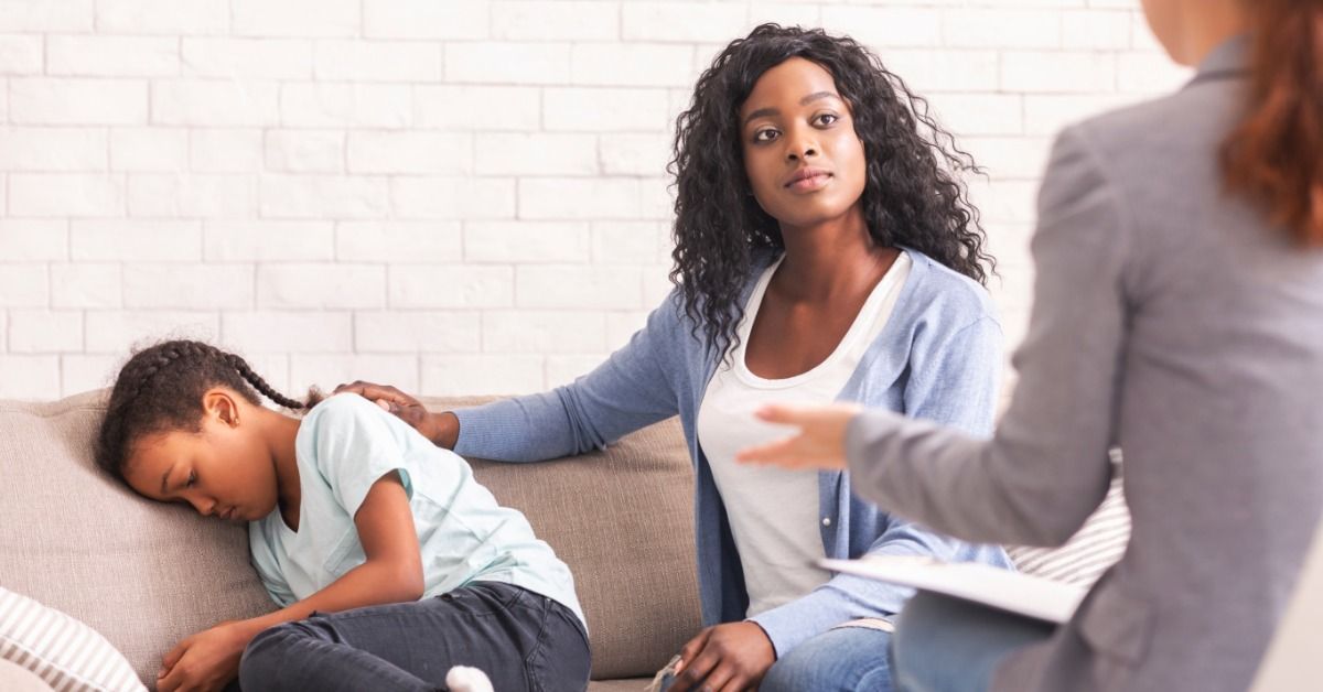A mother comforts her daughter on a sofa while a therapist speaks with them during a therapy session