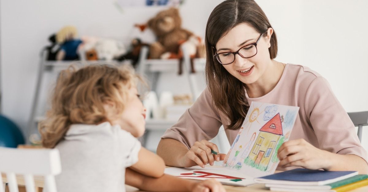 A therapist shows a colorful house drawing to a young child seated across from her at a therapy table.